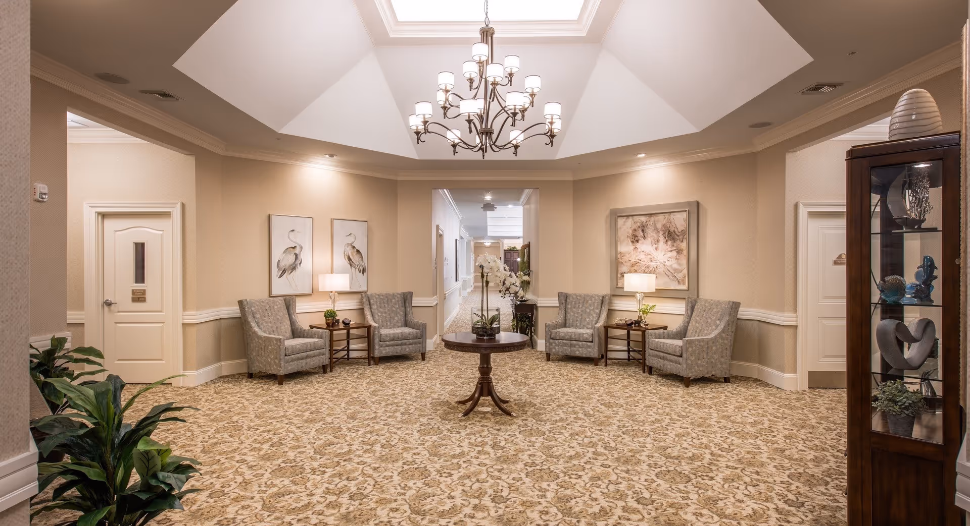 A well-lit, elegant sitting area in a senior living facility with a patterned carpet, four upholstered armchairs arranged in pairs on either side, two side tables with lamps, framed artwork on the walls, a round wooden table with a floral arrangement in the center, a chandelier hanging from a skylight ceiling, and a glass display cabinet with decorative items on the right.