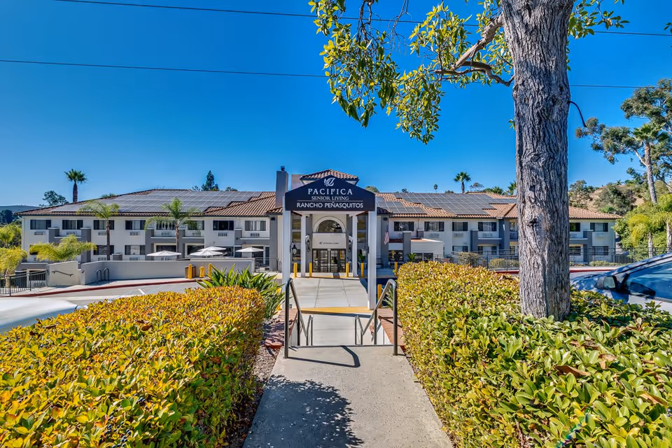 Exterior front view of the Pacifica Senior Living Rancho Penasquitos building with a clear blue sky, landscaped bushes, a tree, and a walkway leading to the entrance.
