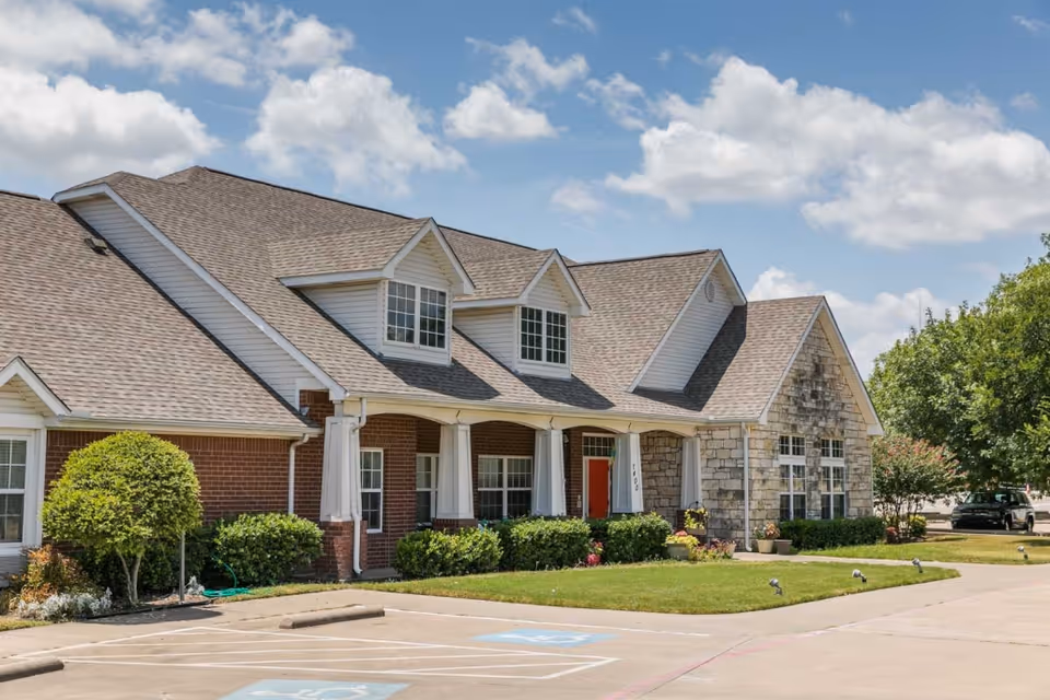 Exterior view of a single-story senior living facility building with a combination of brick and stone facade, multiple windows, a covered porch with white columns, and a red front door. The building is surrounded by neatly trimmed bushes and a well-maintained lawn under a partly cloudy sky. There are parking spaces, including handicapped spots, in front of the building.