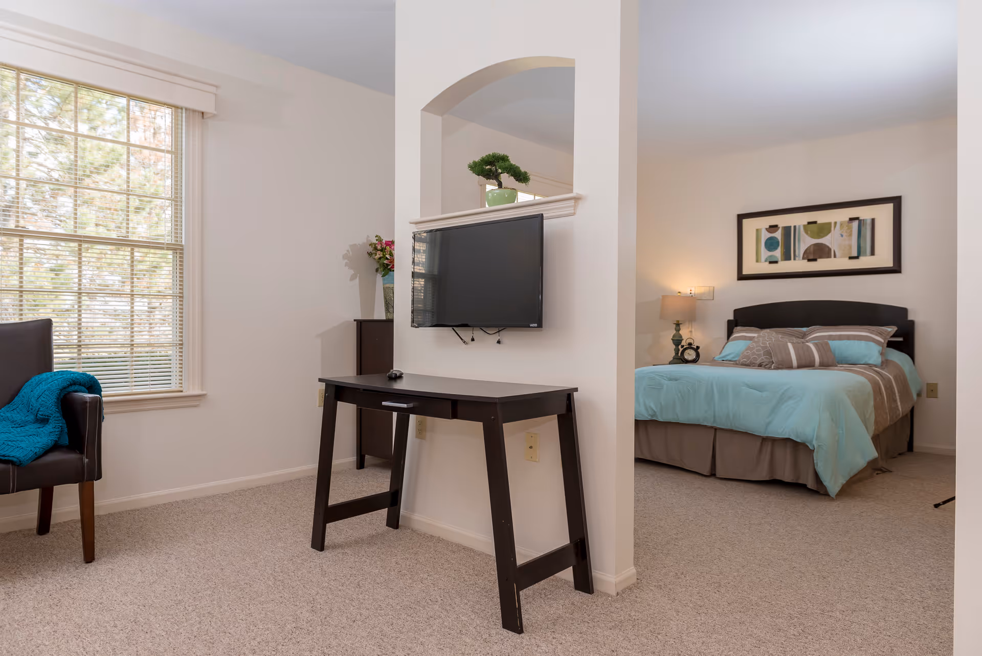 Bright bedroom suite with a bed dressed in teal linens, a wall-mounted TV above a small desk, and a chair by a large window.