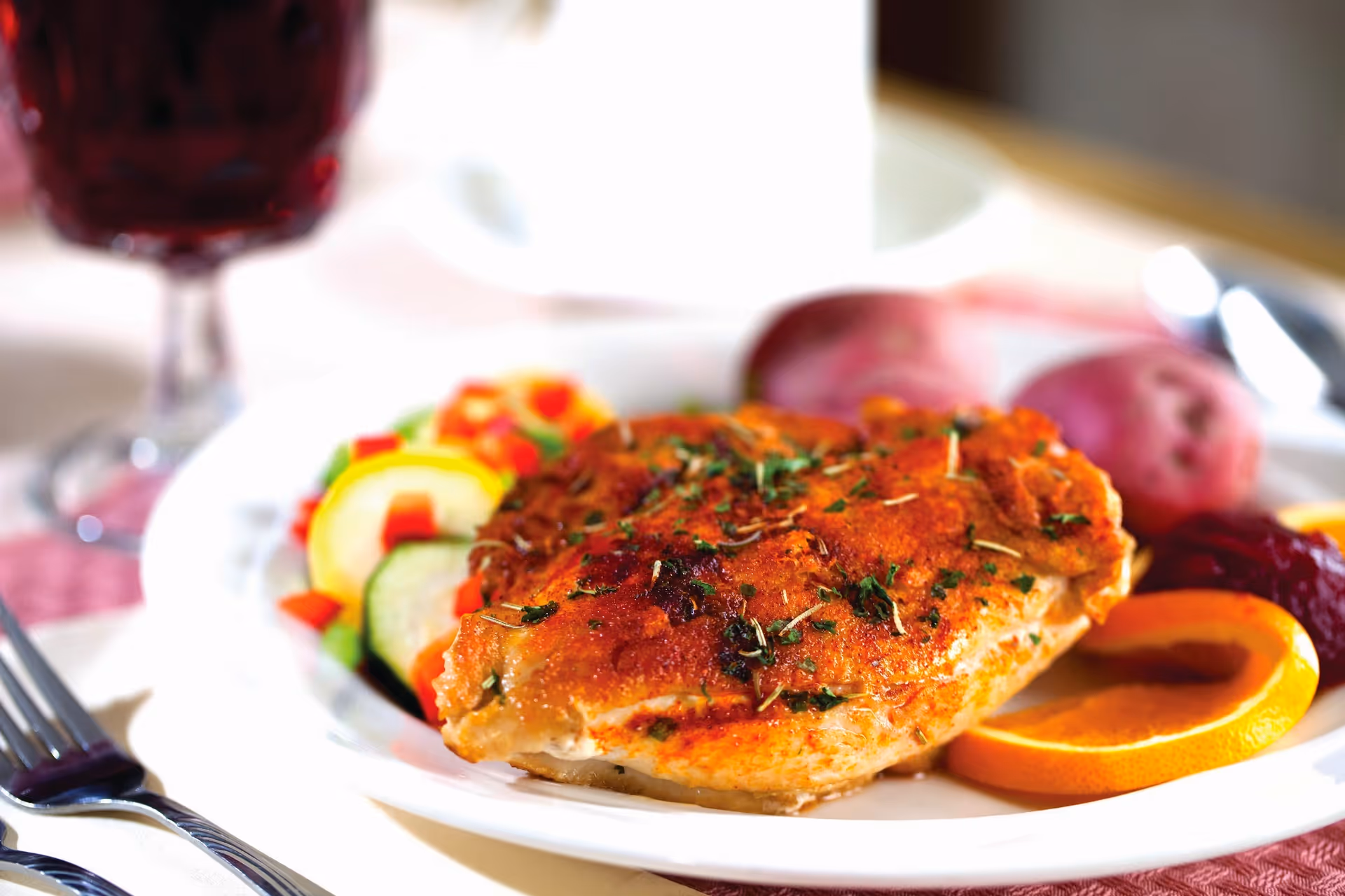 Plated seasoned chicken breast with vegetables, an orange slice and potatoes on a dining table with a glass in the background.