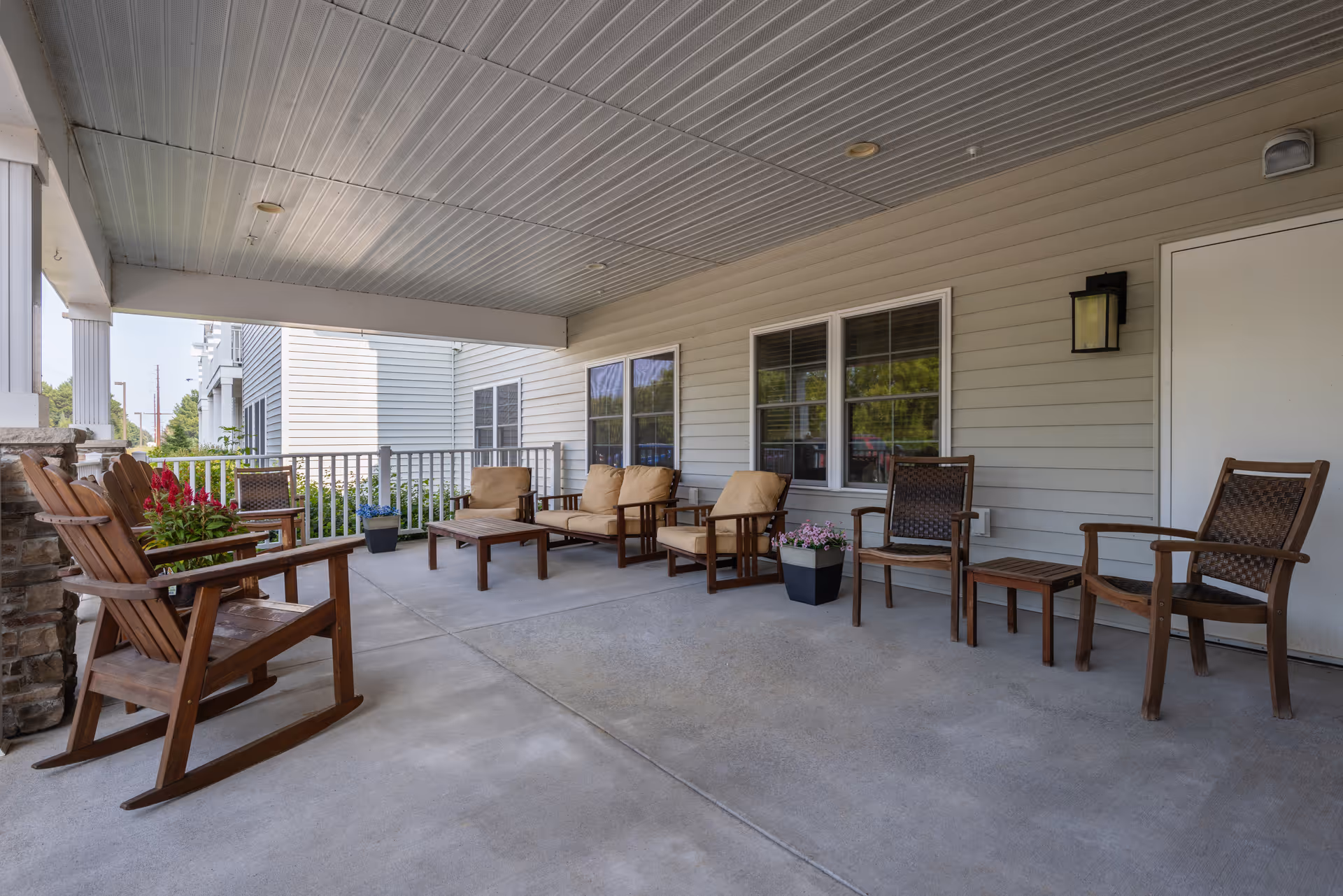 Covered outdoor patio area with wooden rocking chairs, cushioned armchairs, small tables, potted plants, and a railing overlooking greenery. The patio is attached to a building with beige siding and windows.