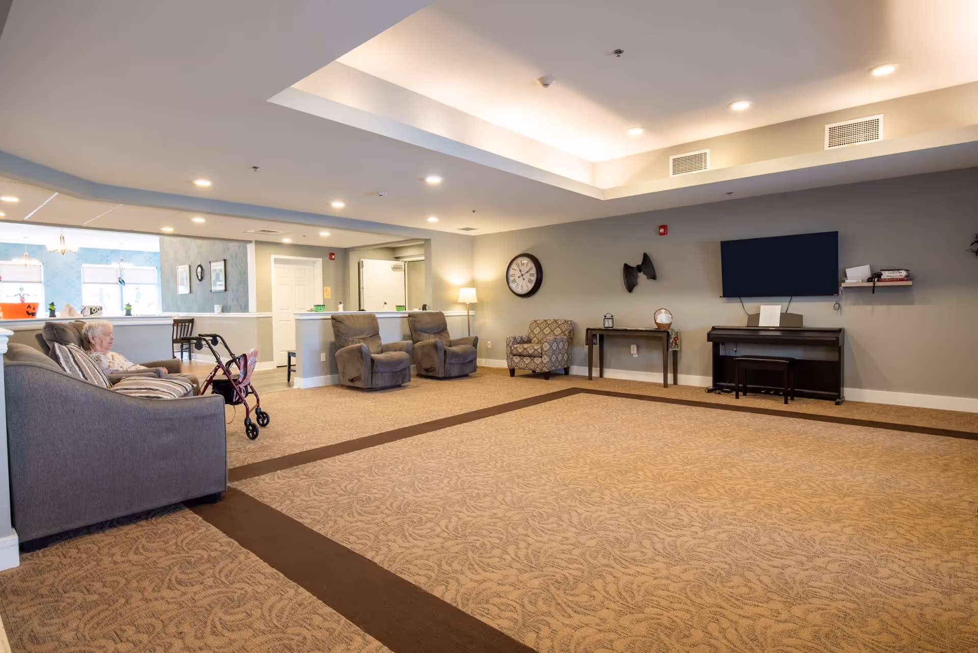 Spacious senior living common lounge with sofas, recliners, a wall-mounted TV, and an elderly resident seated near a walker.