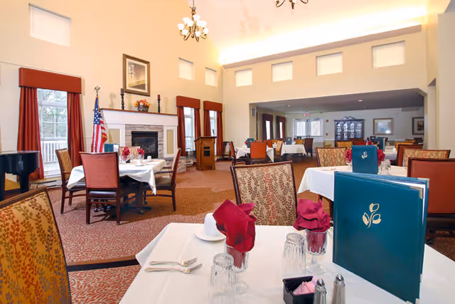A spacious dining room with multiple tables covered in white tablecloths, each set with glasses, silverware, and folded red napkins. The room features high ceilings with small square windows near the top, a fireplace with an American flag beside it, and warm lighting. Chairs have patterned upholstery, and menus are placed on some tables.