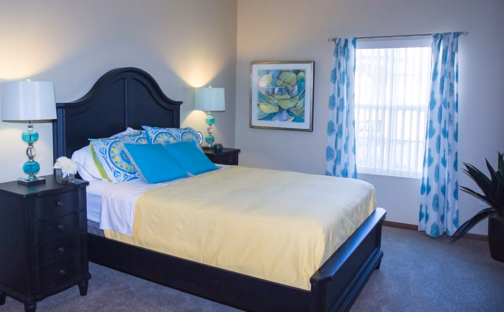 A neatly made bed with a dark wooden headboard and frame, covered with a yellow bedspread and blue decorative pillows. There are two matching bedside tables with turquoise and silver lamps. A framed floral artwork hangs on the wall next to a window with blue patterned curtains. A potted plant is visible in the corner of the room.