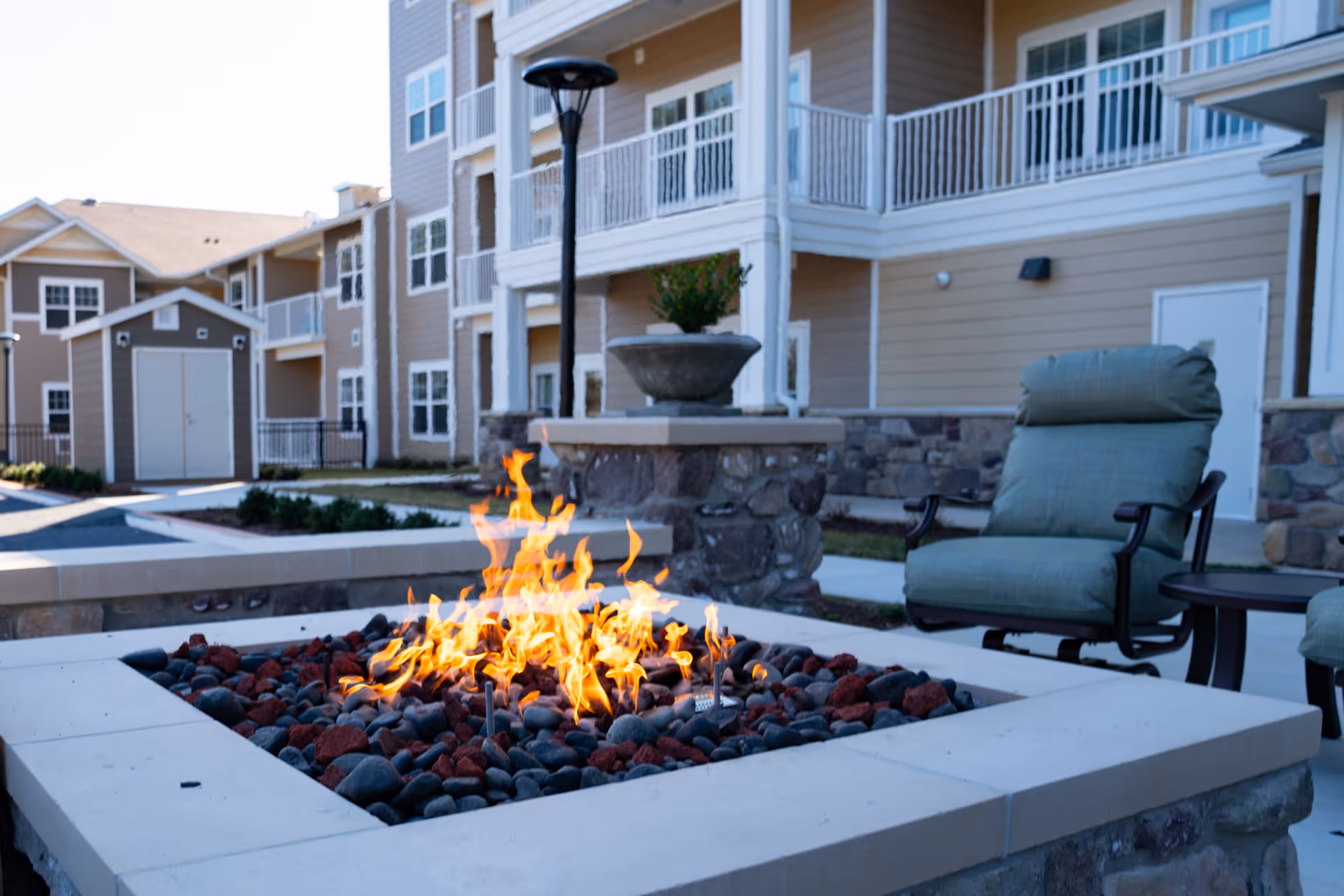 Outdoor seating area at Wilshire Estates Gracious Retirement Community featuring a stone fire pit with flames surrounded by black and red stones, cushioned chairs, and a multi-story beige building with white railings in the background.