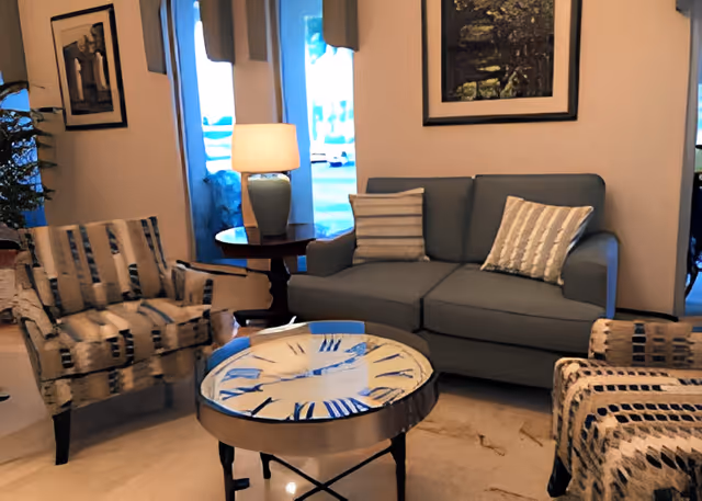 A cozy living room area with a gray sofa adorned with two striped pillows, two patterned armchairs, a round coffee table with a clock face design, a side table with a lamp, and framed artwork on the walls.