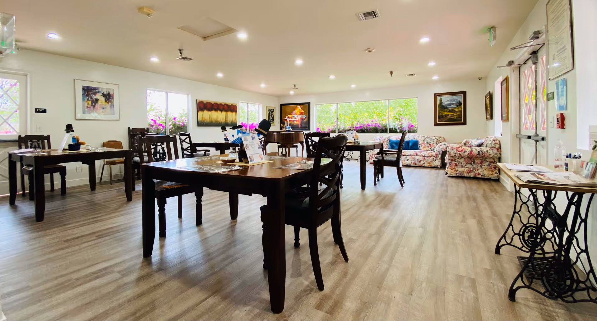 A bright and spacious common area with wooden tables and chairs arranged on a light wood floor. The room has large windows with flower boxes outside, letting in natural light. There are floral-patterned sofas along the far wall, colorful paintings on the walls, and a piano in the corner. The space appears welcoming and comfortable, suitable for socializing or activities.