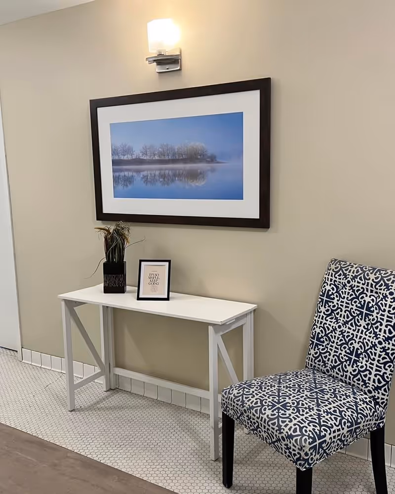 A small white table against a beige wall with a decorative plant and a framed quote on top. Above the table is a large framed landscape photograph of trees reflected in calm water under a blue sky. To the right of the table is a chair with a blue and white patterned upholstery. The floor has small white hexagonal tiles near the wall and wood flooring in the foreground.