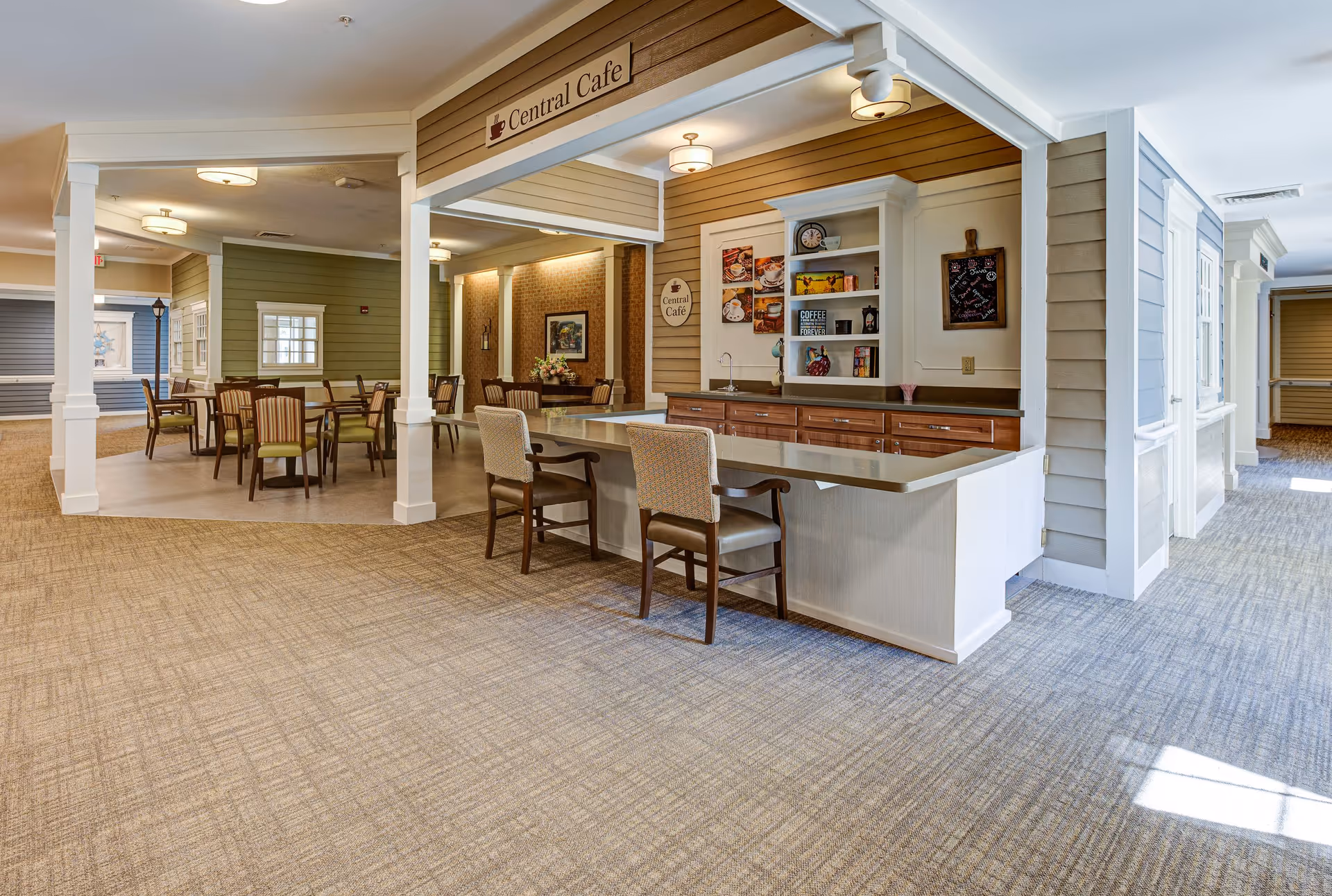 Interior view of a senior living facility's Central Cafe area featuring a counter with two chairs, wooden cabinetry, and shelves with decorative items. In the background, there are multiple tables and chairs arranged for dining, with warm lighting and a cozy atmosphere.