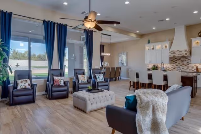 A spacious and modern living area featuring four dark leather recliners with decorative pillows, a tufted ottoman in the center, a gray sofa with a white throw blanket, and large windows with blue curtains letting in natural light. In the background, there is a dining area with a table and chairs, and a kitchen with white cabinetry, a tiled backsplash, and a large island with bar stools.