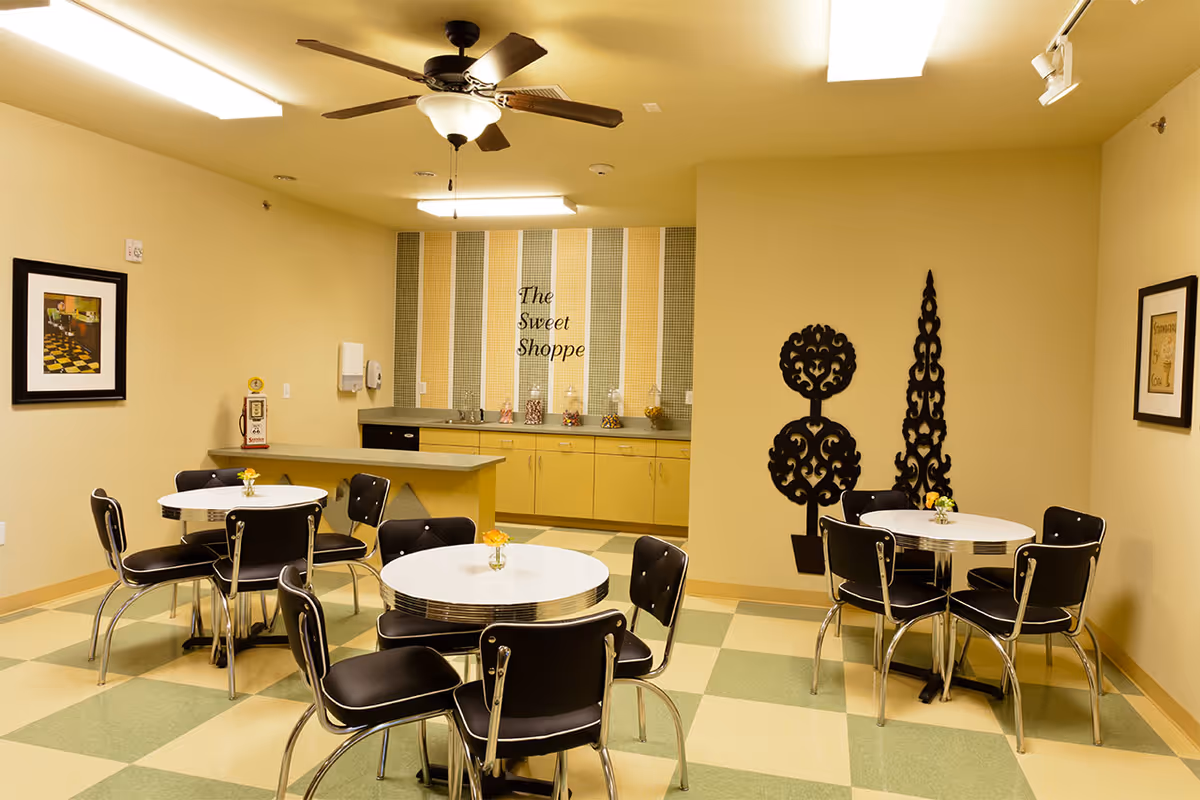 A small cafe-style dining room with round retro tables and black chairs in front of a counter labeled "The Sweet Shoppe".