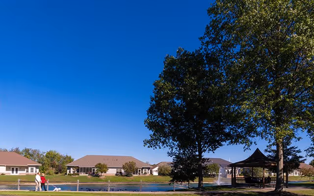 Pond and gazebo on a senior living campus with single-story buildings, a fountain, large trees, and two people walking a dog under a clear blue sky.