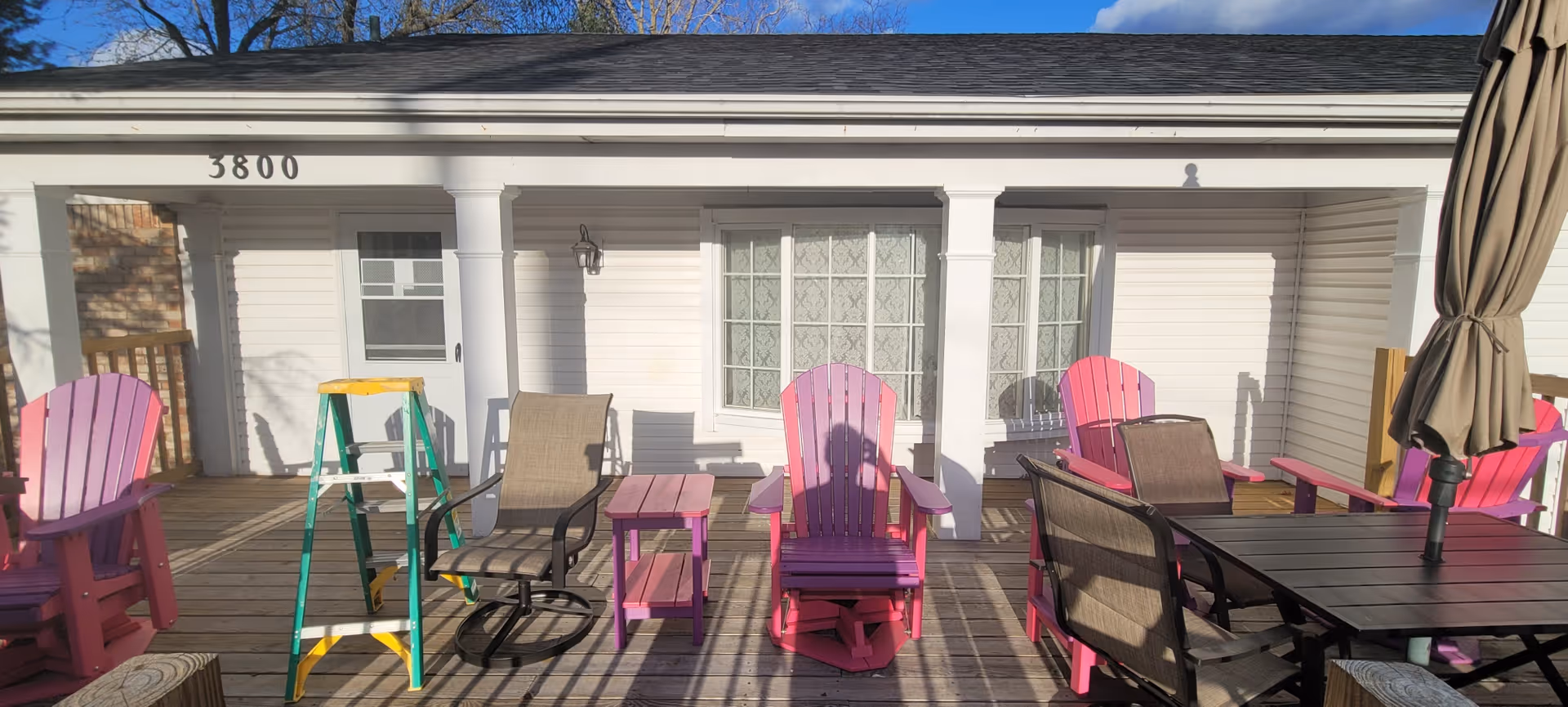 Outdoor wooden deck area with several pink Adirondack chairs, a small pink side table, a brown patio table with matching chairs, a closed beige patio umbrella, and a green and yellow step ladder. The deck is attached to a white building with the number 3800 above the door and windows with lace curtains.