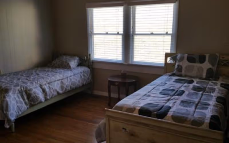 A simple bedroom with two single beds placed against opposite walls. Each bed has patterned bedding and a pillow. Between the beds is a small round wooden table. A large window with blinds is centered on the wall behind the table, allowing natural light into the room. The floor is wooden, and the walls are painted a neutral color.