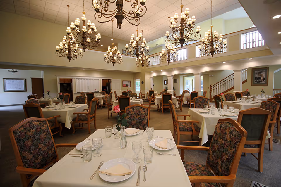 Spacious dining room with multiple set tables, floral-upholstered chairs, and ornate chandeliers in a retirement community.