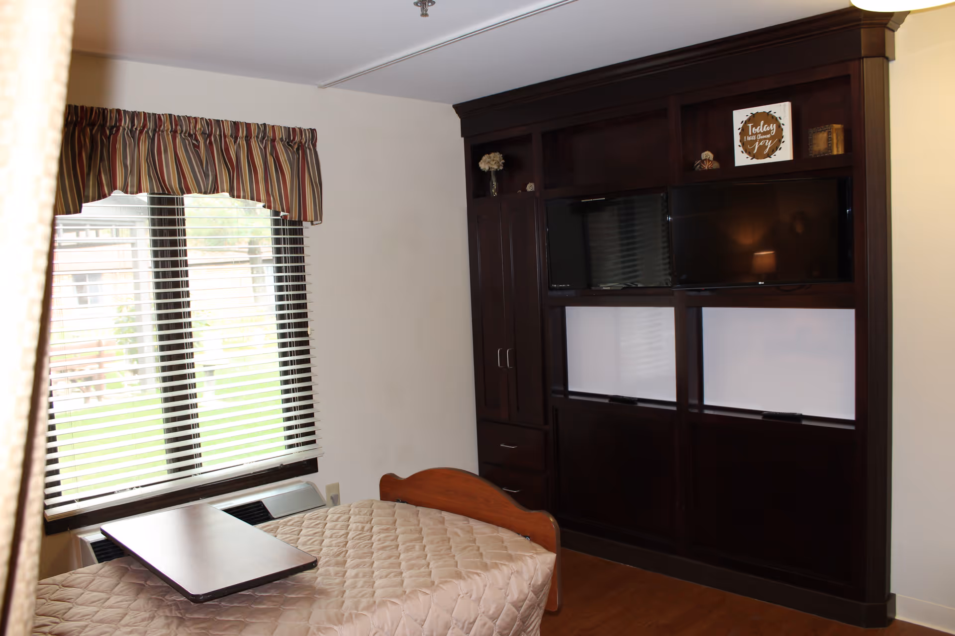A bedroom in a senior living facility featuring a single bed with a beige quilted bedspread, a dark wooden entertainment unit with two televisions and decorative items, a window with striped valance and blinds, and a small overbed table.