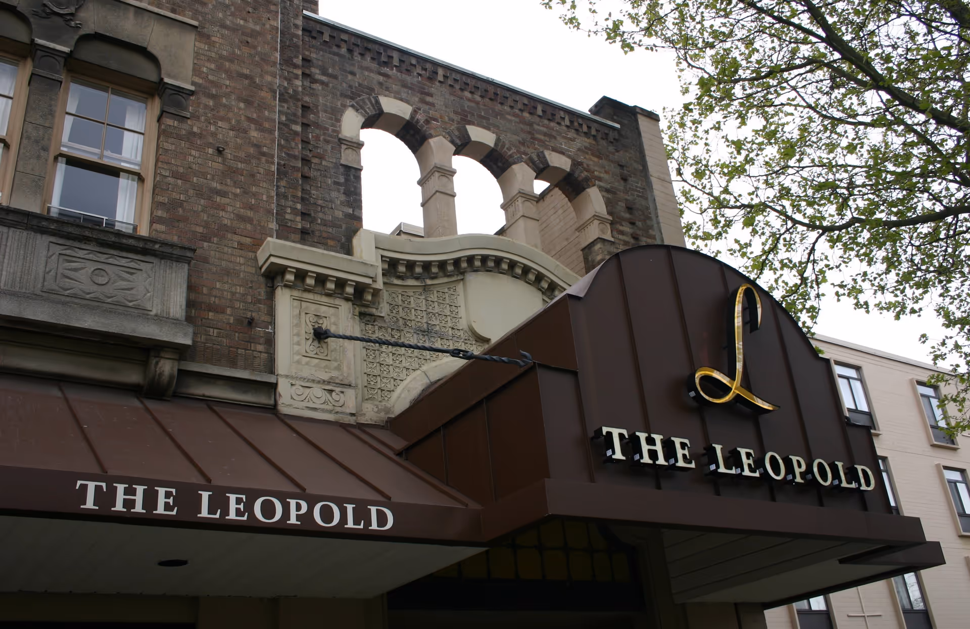 Exterior view of The Leopold Retirement Residence showing a brown awning with the name 'THE LEOPOLD' in white letters and a decorative building facade with arched windows and brickwork.