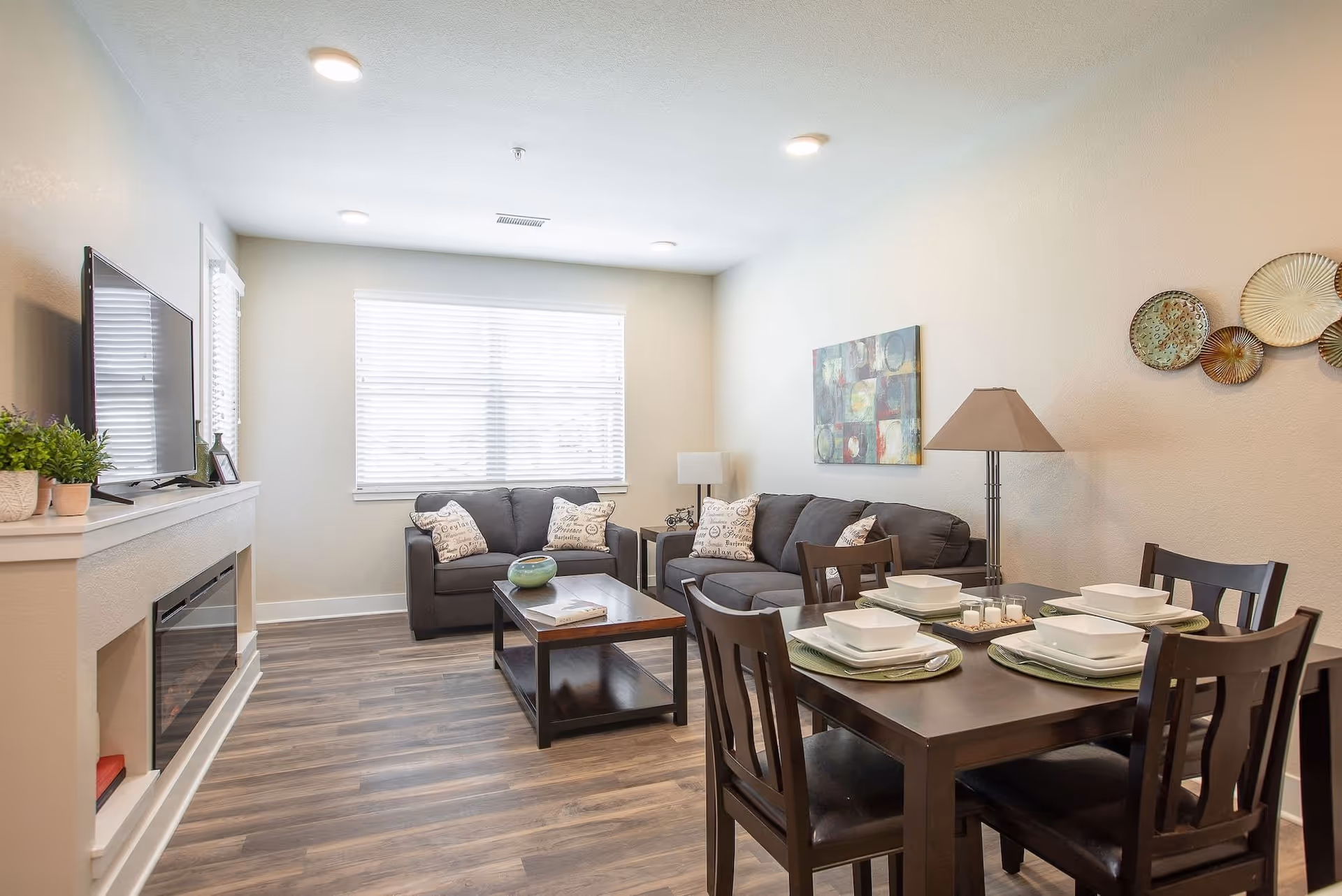 A bright living and dining area with two gray sofas, a wooden coffee table, a dining table set for four with white square dishes, a floor lamp, wall art, and a TV mounted above a modern electric fireplace. The room has wood flooring and a large window with white blinds.