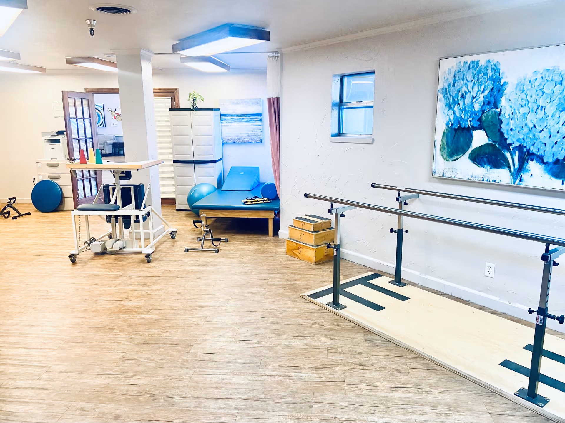 A rehabilitation room with parallel bars for walking exercises, a blue padded therapy table, exercise balls, wooden step boxes, and various physical therapy equipment. The room has light-colored walls and flooring, a small window, and a large painting of blue flowers on the wall.
