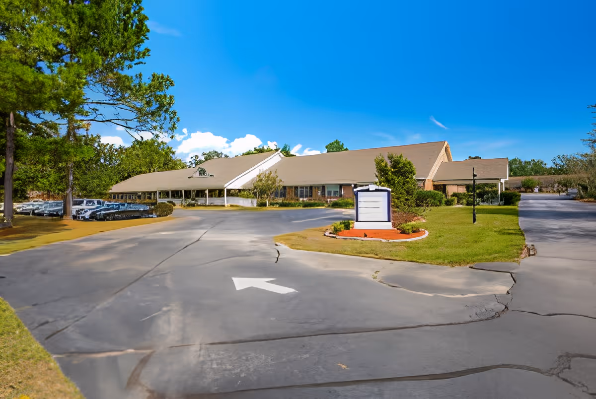 Single-story senior living facility building with a driveway, landscaped sign island and parked cars under a blue sky.