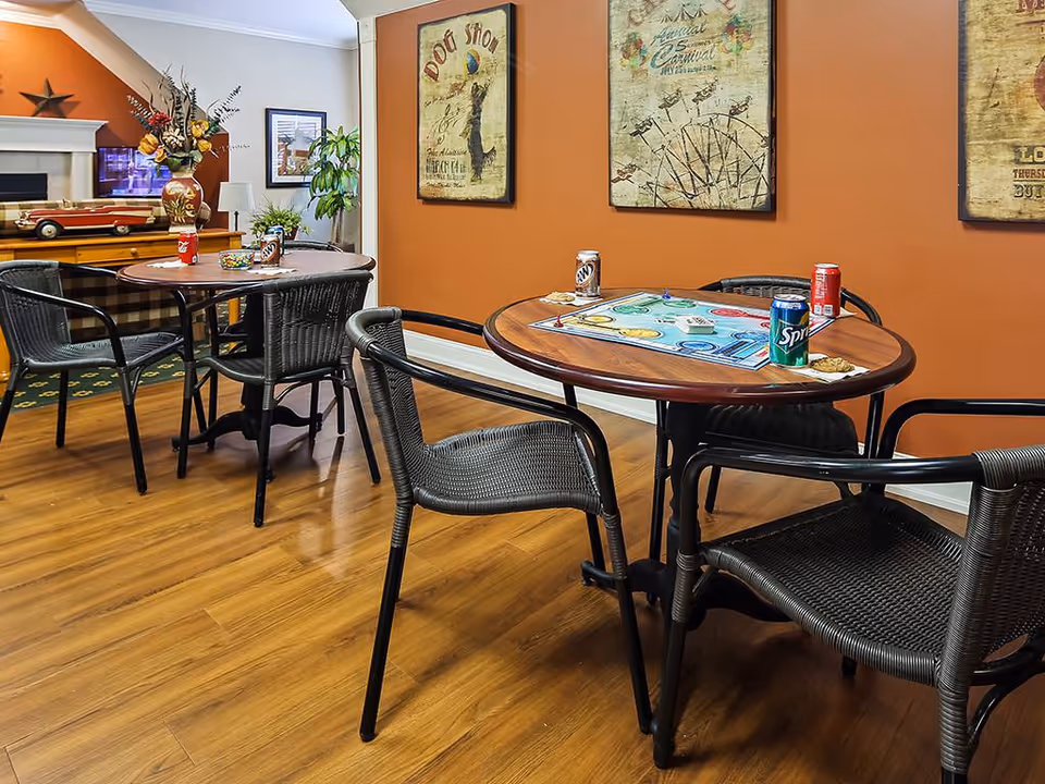 A cozy common area with two round wooden tables surrounded by black wicker chairs. One table has a board game set up with soda cans and cookies, while the other table has a vase with flowers and a model car. The room has warm orange walls decorated with vintage-style framed posters, and a wooden floor.