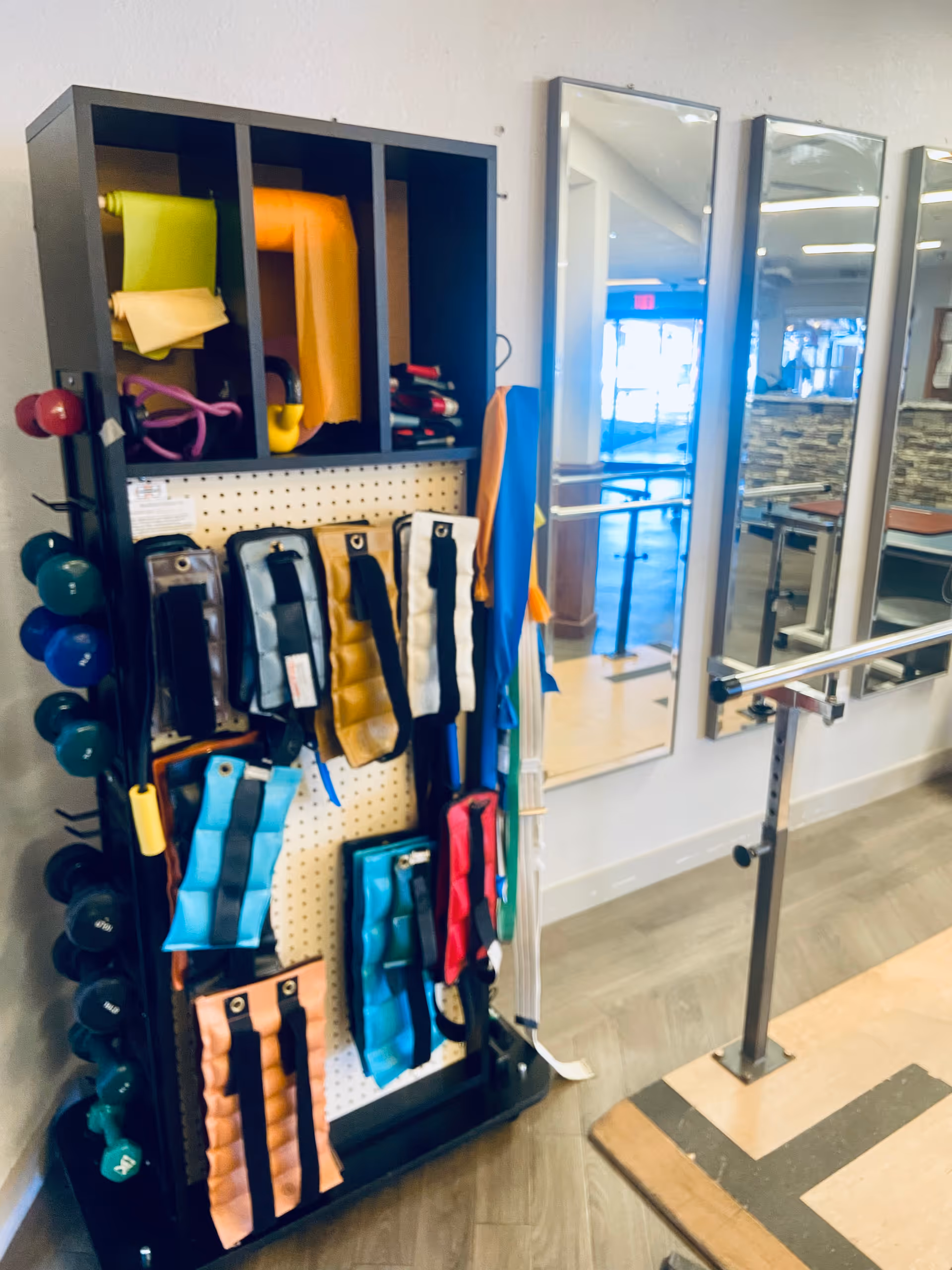 A fitness equipment rack holding various exercise bands, weights, and resistance tools next to a wall with three large mirrors and a ballet barre in a rehabilitation or exercise room.