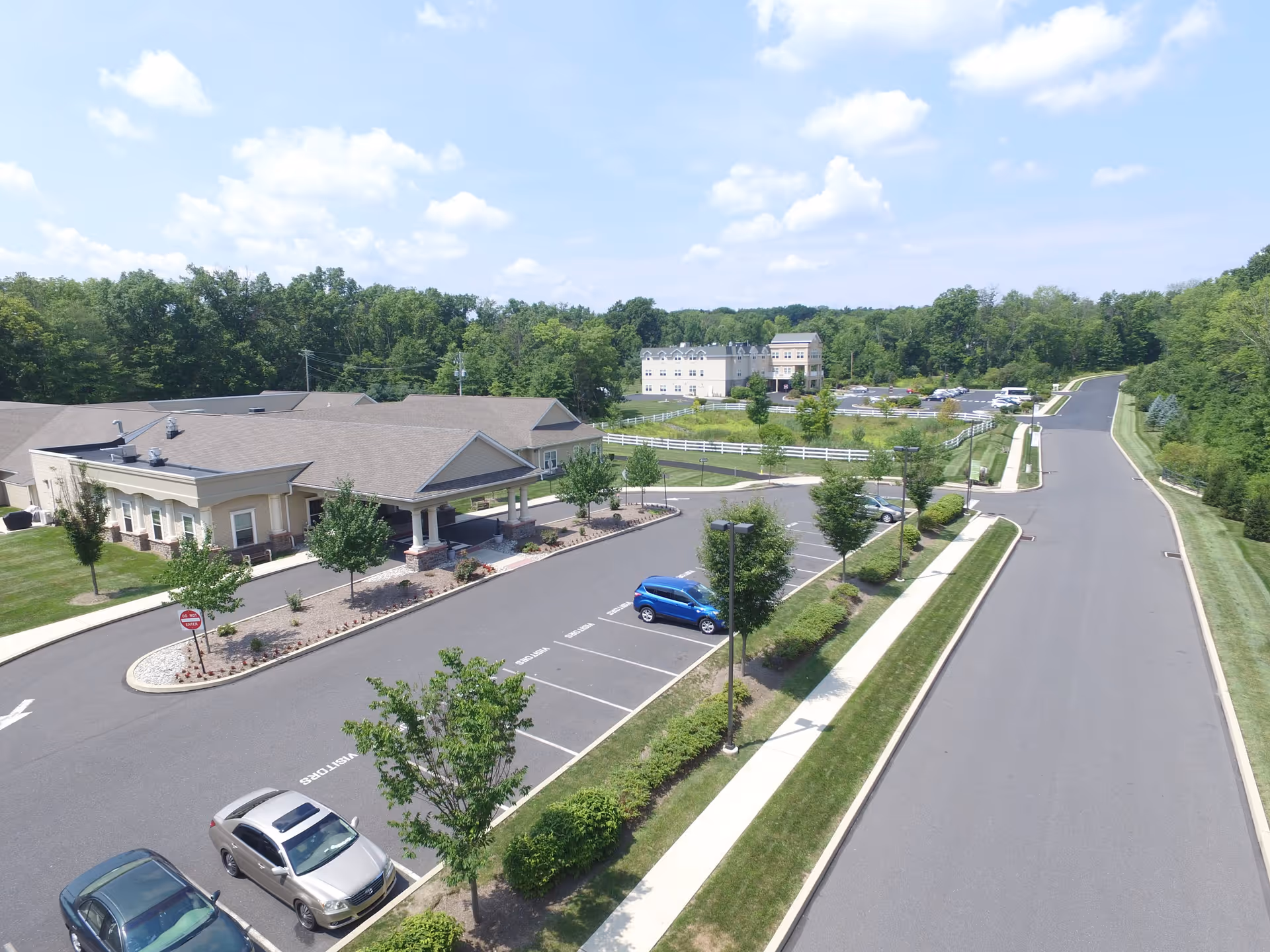 Aerial view of Hidden Meadows On the Ridge facility showing a large building with a covered entrance, a parking lot with several cars, surrounding greenery, trees, and a road leading away from the facility under a partly cloudy sky.