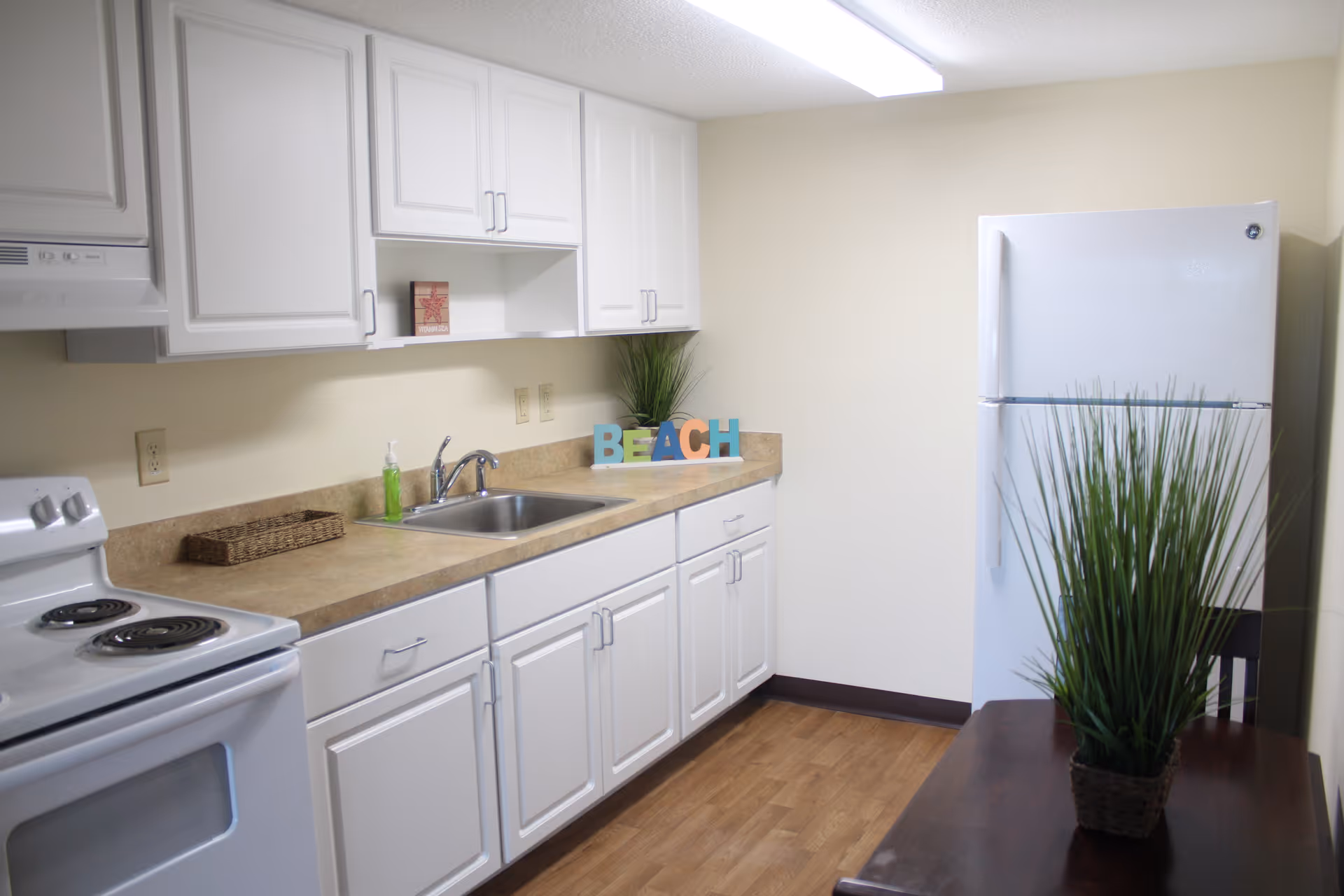 A clean kitchen with white cabinets, a white electric stove, a stainless steel sink, and a white refrigerator. The countertop has a small green soap dispenser, a wicker tray, and decorative letters spelling 'BEACH' next to a potted plant. There is also a tall potted plant on a dark wooden table in the foreground.
