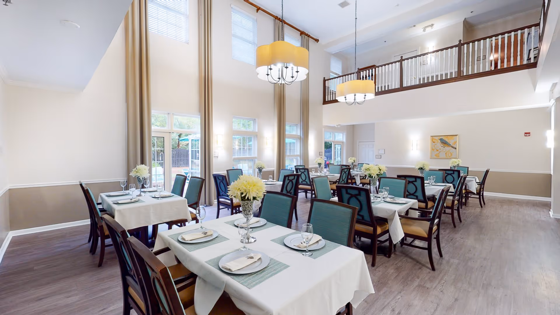 A bright and spacious dining room with multiple tables covered in white tablecloths and set with plates, napkins, and glasses. Each table has a centerpiece of white flowers in a vase. The room features high ceilings with large windows letting in natural light, beige walls with white trim, and two large hanging light fixtures. There is a balcony railing visible on the upper level and a door leading outside.