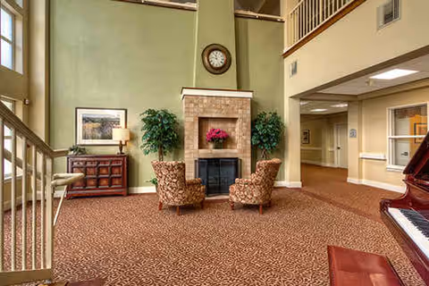 Spacious senior living common room with a central stone fireplace flanked by two armchairs, a piano, and high ceilings.