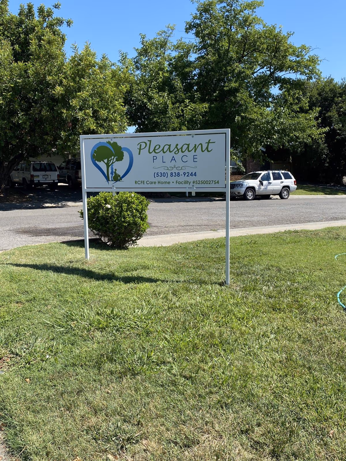 A roadside sign for "Pleasant Place" on a grassy lawn with trees and parked cars behind it.