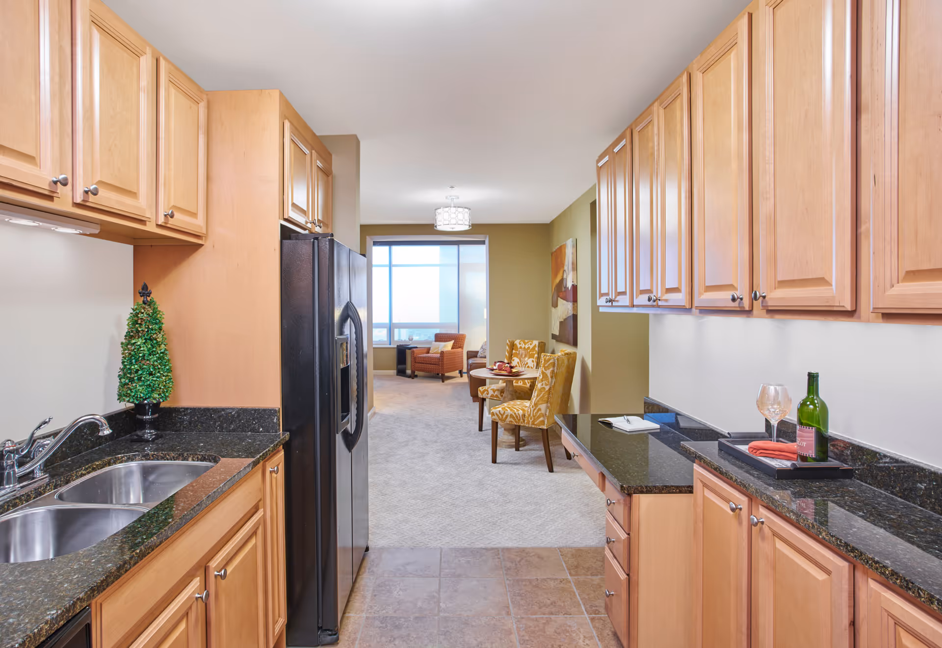 A galley kitchen with light wood cabinets, black granite countertops and a stainless refrigerator opening into a carpeted sitting area with chairs.