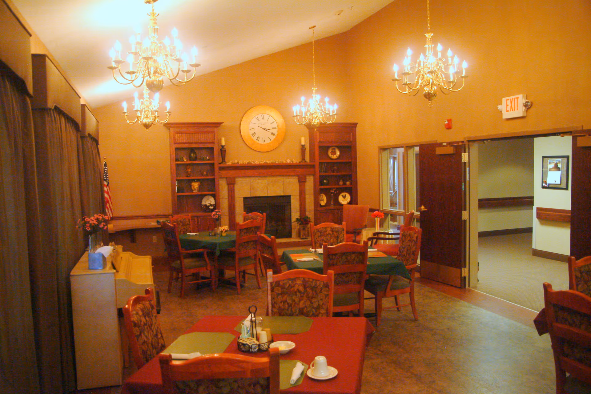 Communal dining room with multiple set tables, chandeliers, a fireplace with a clock, and an exit doorway.