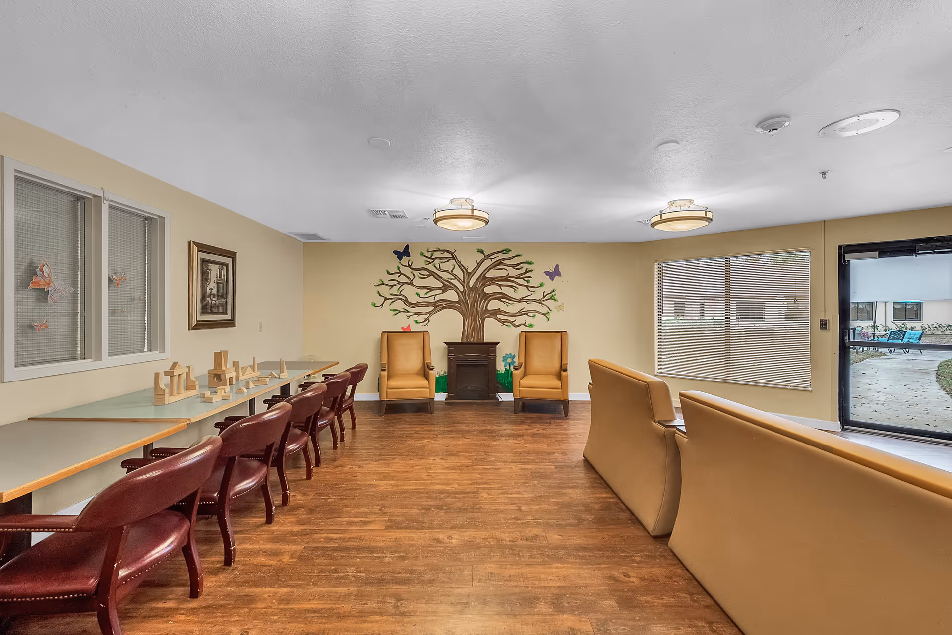 A common area in a senior living facility with wooden flooring, beige walls, and ceiling lights. There are two mustard-colored armchairs and a small cabinet against the far wall, which has a painted tree with butterflies and flowers. To the left, there is a long table with several red chairs and wooden block toys on top. On the right side, there are beige sofas facing away from the camera, and a large window and glass door leading outside.