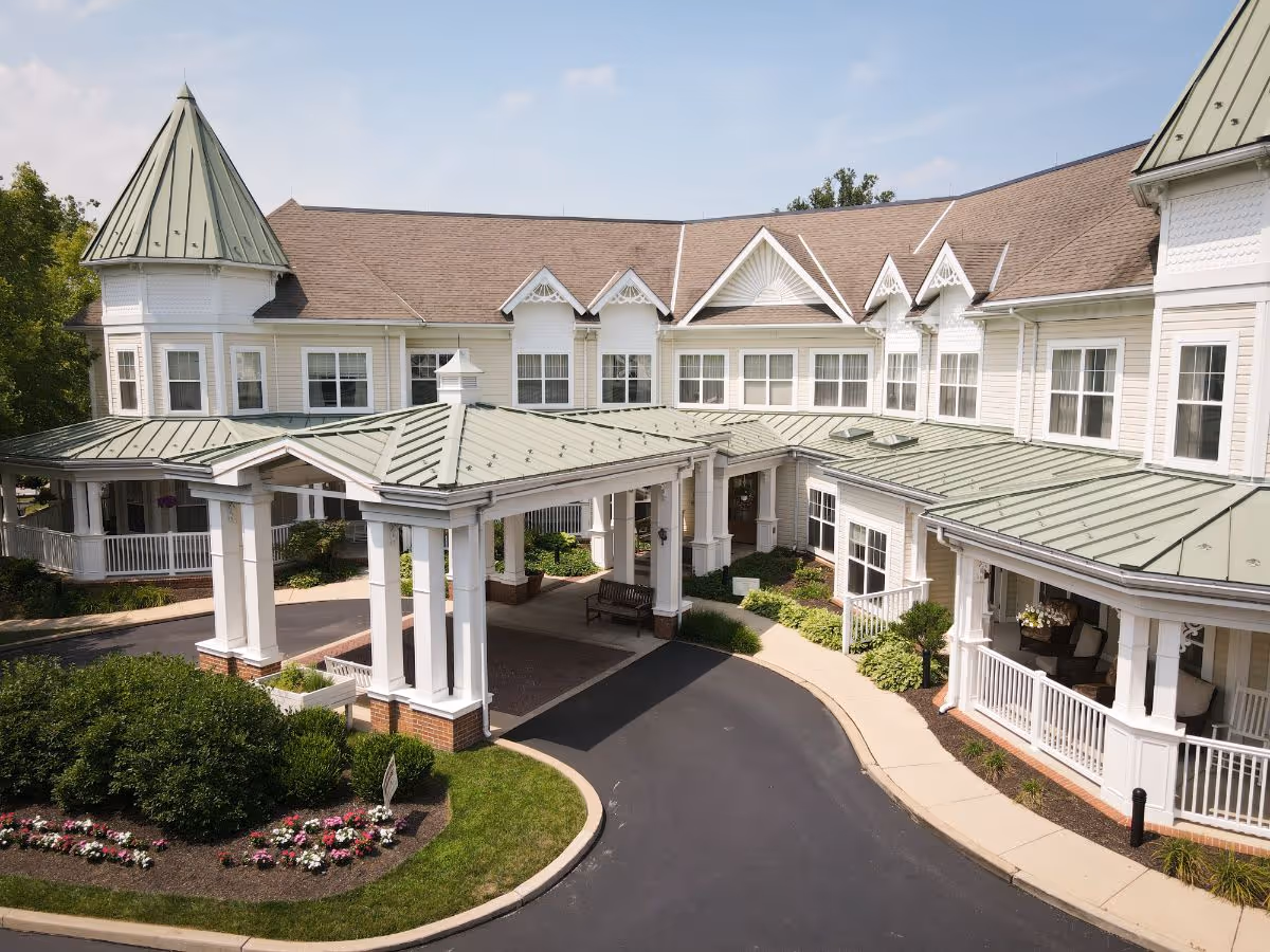 Exterior view of Sunrise of Granite Run, a senior living facility with a large covered entrance, white columns, green metal roofs, and beige siding. The building has multiple windows and decorative architectural details, surrounded by landscaped bushes and flowers.