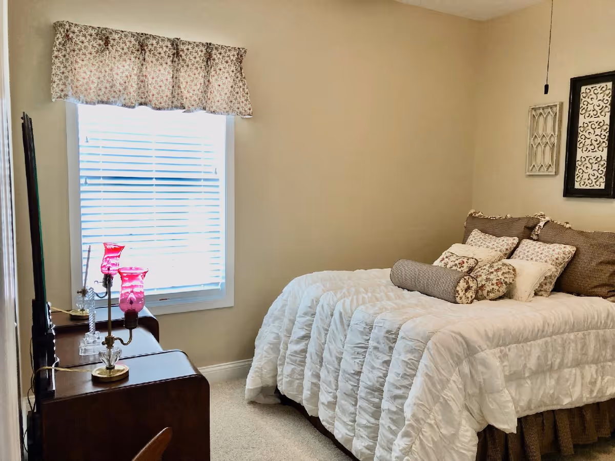 A cozy bedroom with a neatly made bed featuring a white comforter and multiple decorative pillows. There is a window with floral curtains letting in natural light. A wooden dresser with two pink glass lamps is positioned against the wall opposite the bed. The walls are painted beige and decorated with framed artwork.