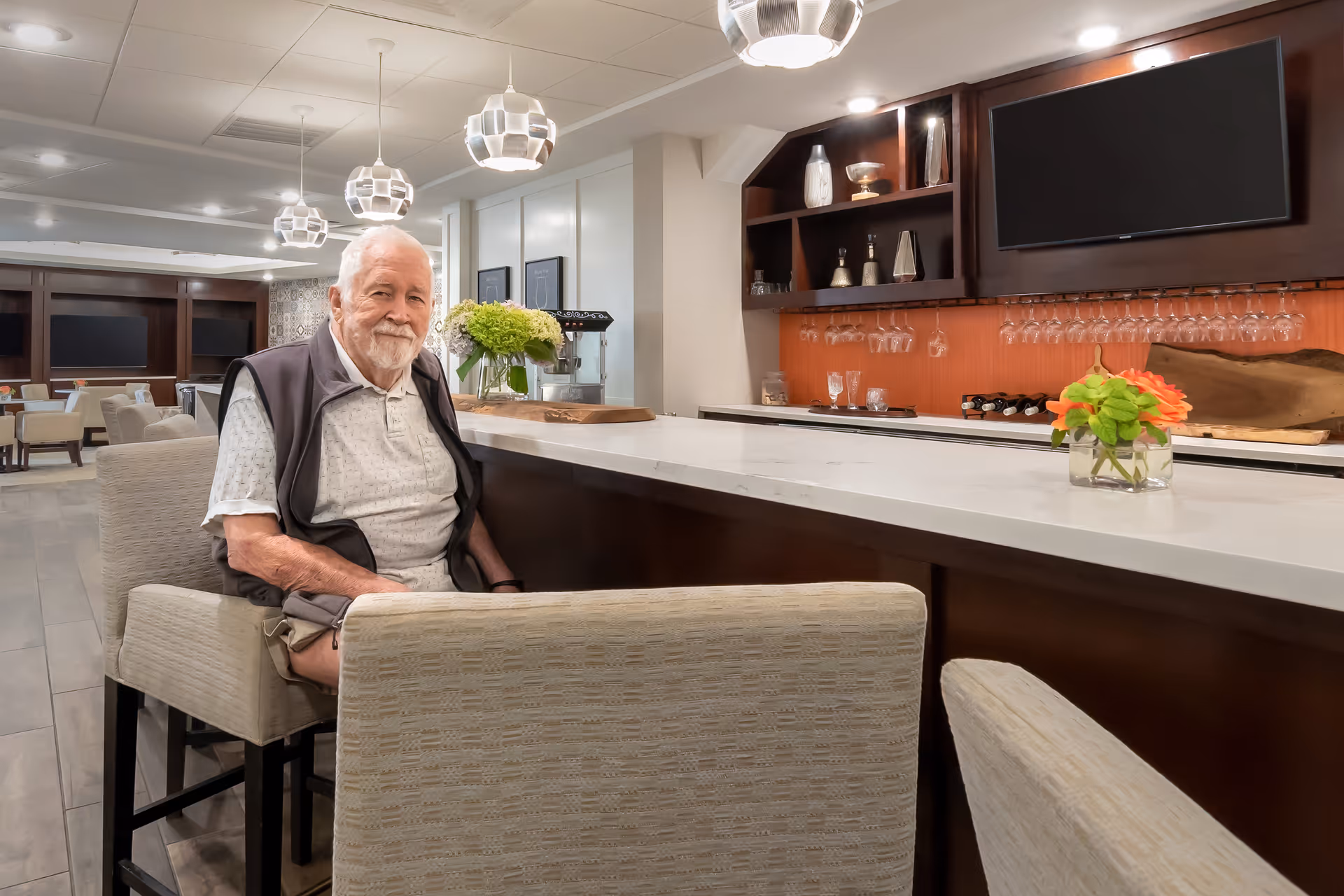 An elderly man with white hair and a beard sits on a cushioned bar stool at a modern bar counter in a senior living facility. The bar area features a white countertop, hanging pendant lights, glassware, and a flat-screen TV mounted on the wall. The background shows a spacious, well-lit common area with tables and chairs.