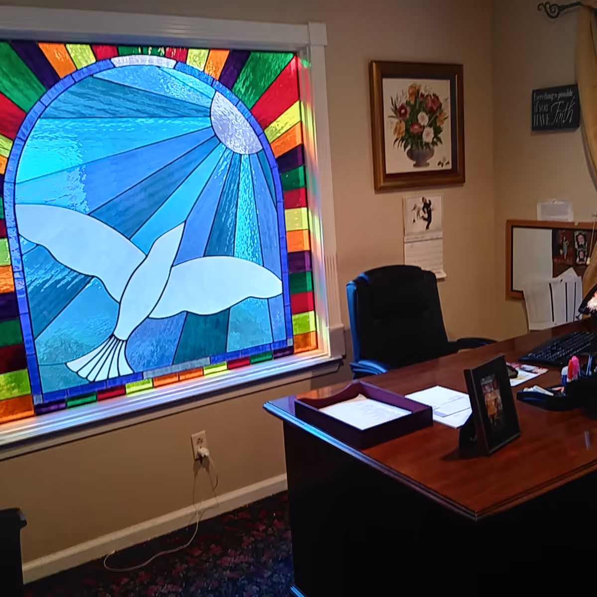 Office interior with a wooden desk and chair beside a large colorful stained-glass window depicting a dove.