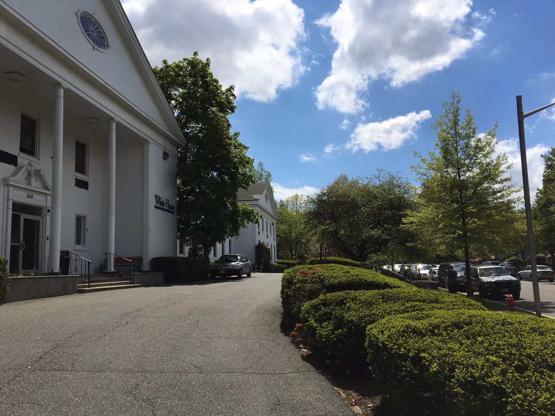 Front exterior of a white healthcare building with large columns, a driveway lined by trimmed hedges and parked cars under a blue sky.