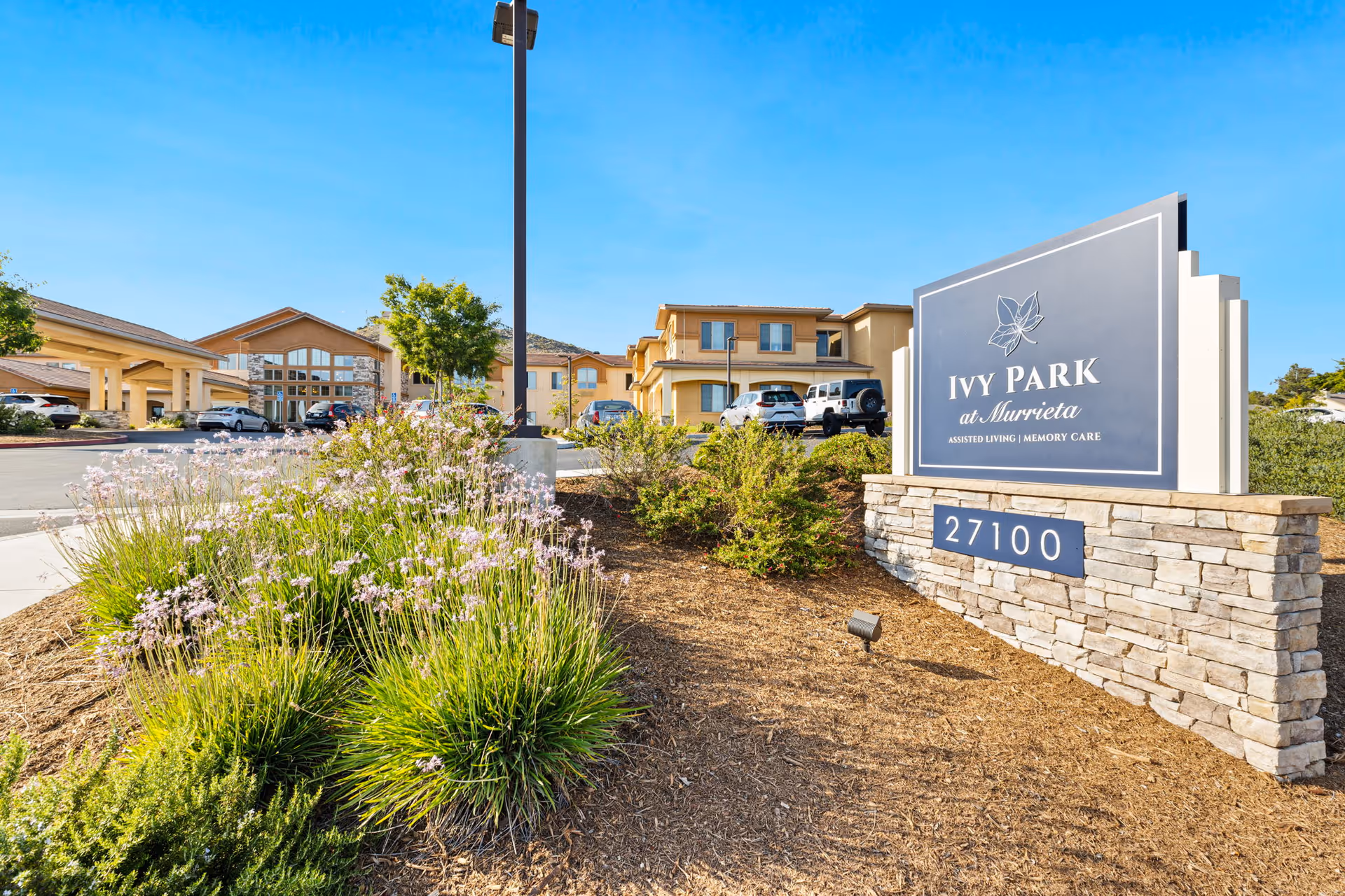 Entrance sign reading 'Ivy Park at Murrieta' beside landscaped beds with the facility building and parking lot in the background under a clear blue sky.