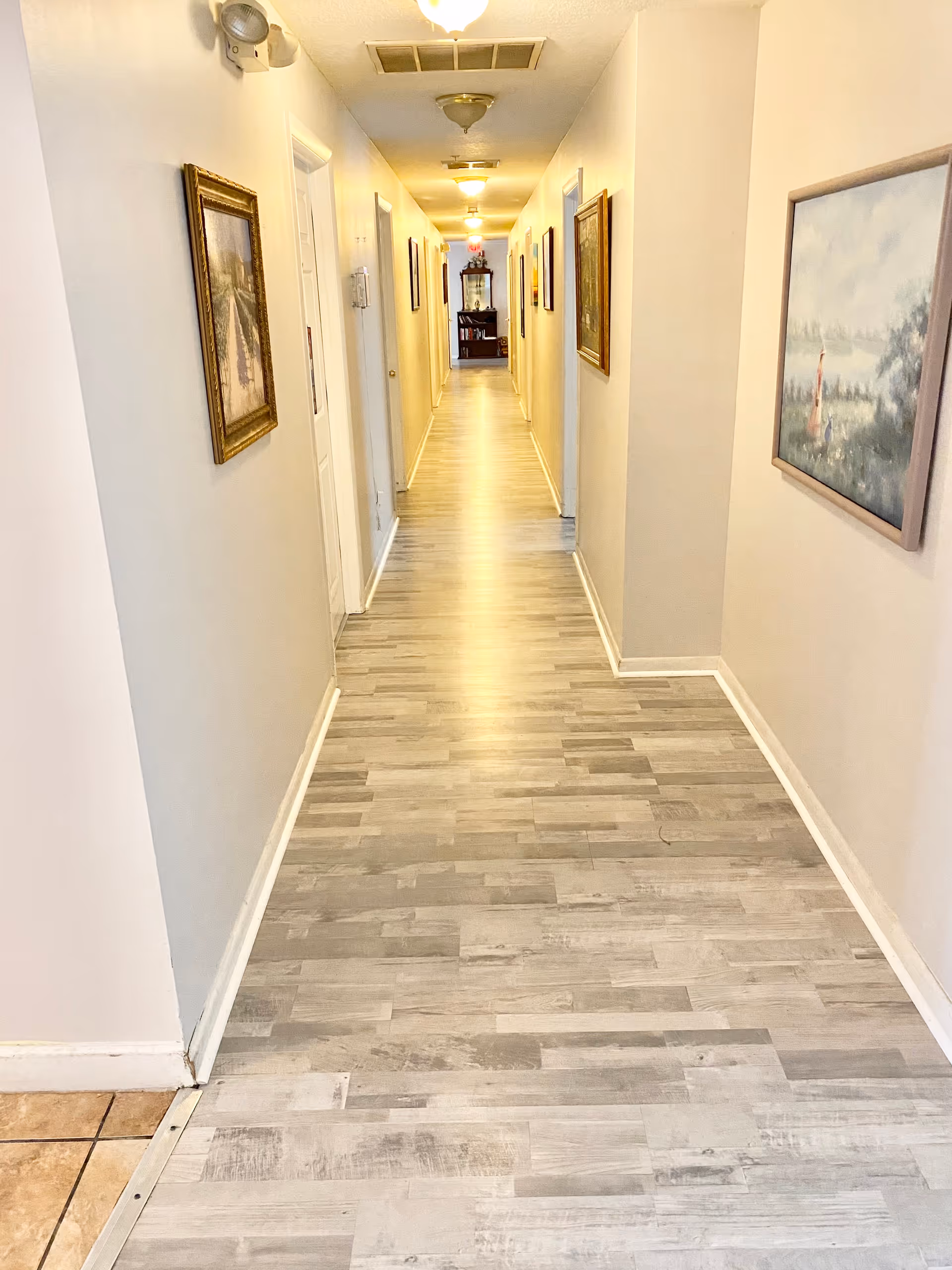 A long, well-lit hallway with light-colored walls and wood-patterned flooring. Several framed paintings hang on both walls, and multiple doors line the corridor. At the far end, there is a piece of furniture with decorative items on top.