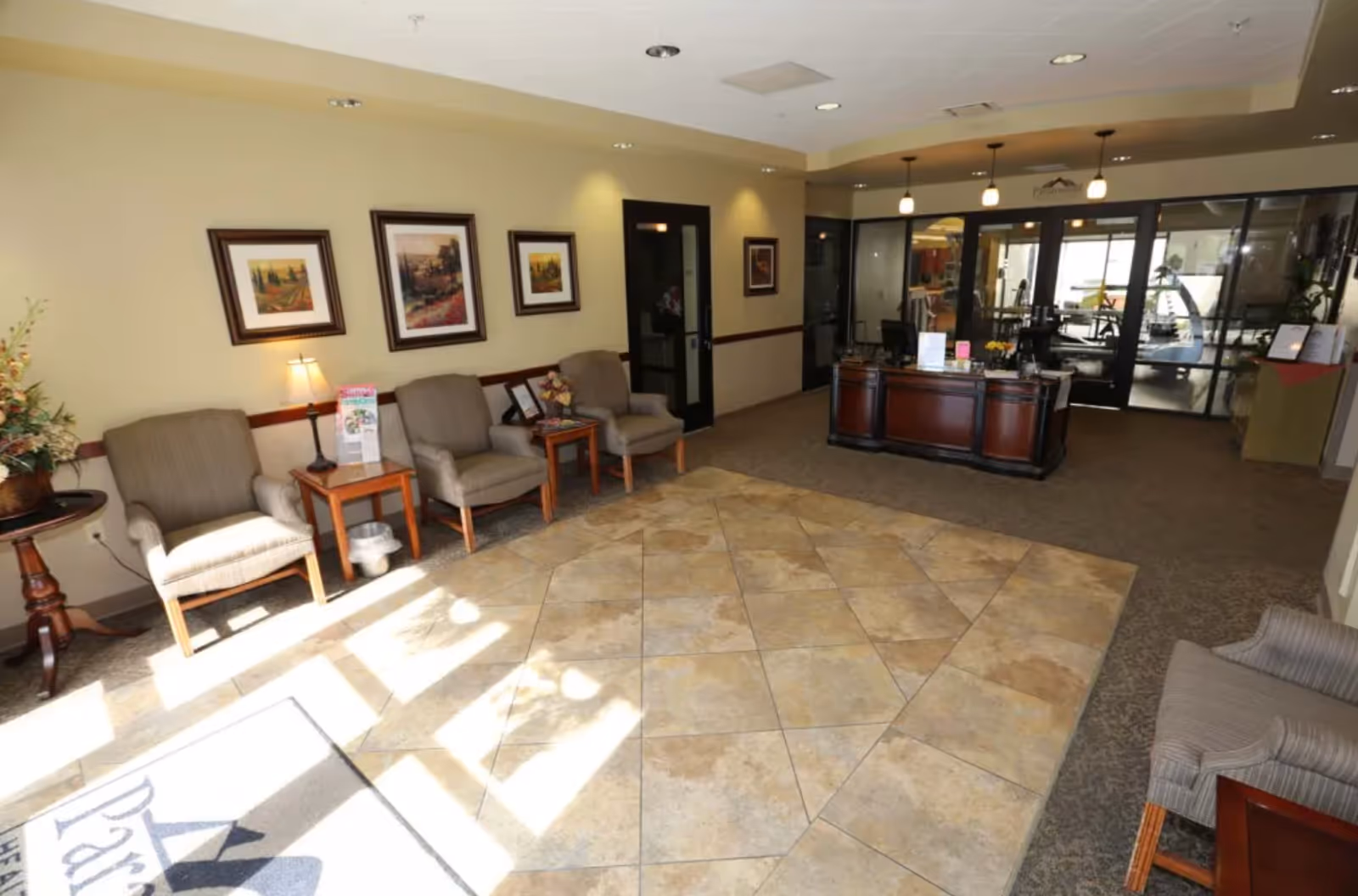 A well-lit reception area with a tiled floor section in the center and carpeted edges. There are four cushioned chairs along the left wall with small tables and a lamp between them. Three framed paintings hang on the wall above the chairs. A wooden reception desk is positioned towards the back, with glass doors behind it leading to another room. A small round table with a flower arrangement is visible on the left side near the chairs.