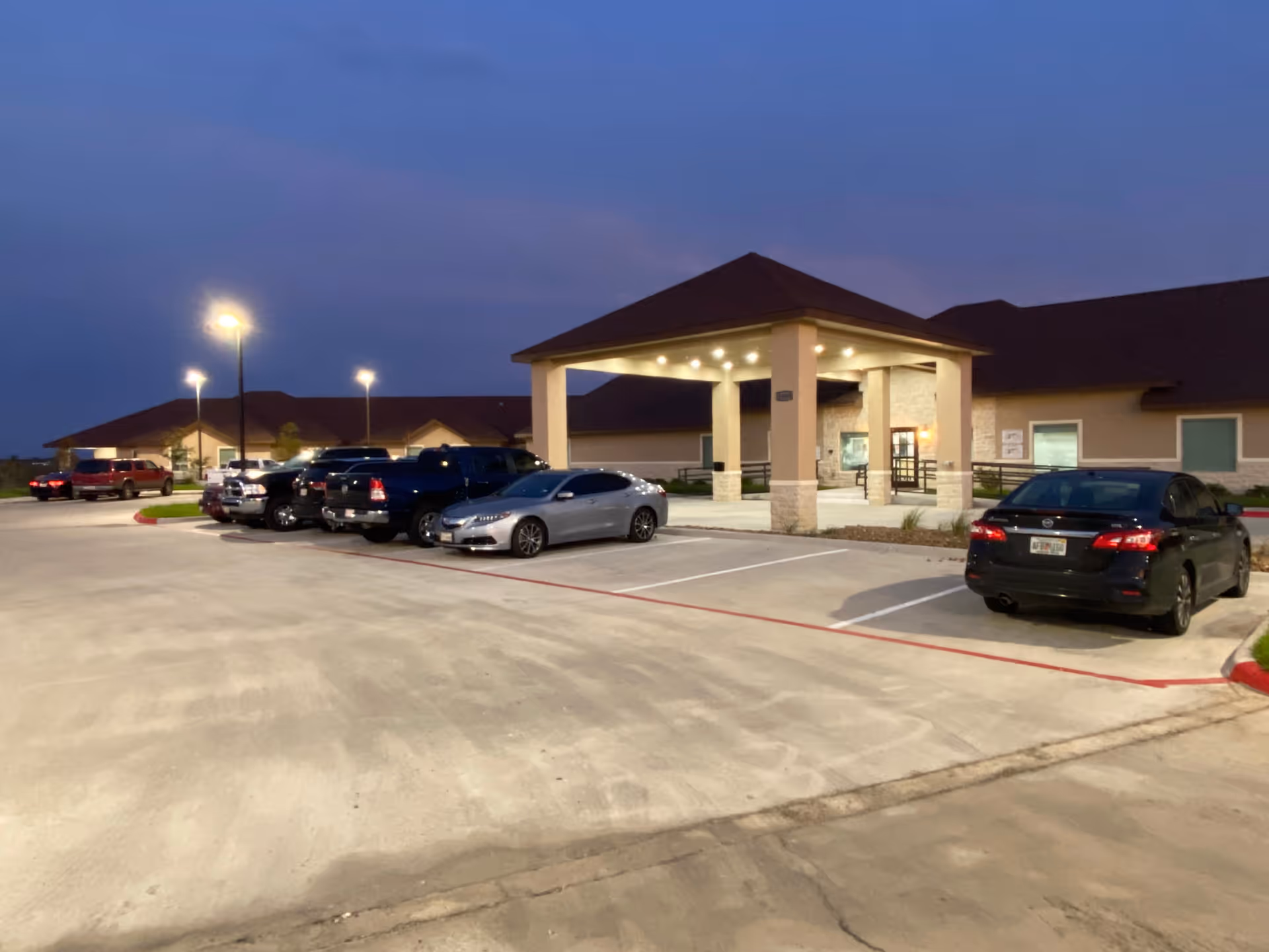 Exterior view of Mont Belvieu Rehabilitation & Healthcare Center at dusk, showing the entrance with a covered drop-off area, several parked cars, and illuminated street lamps.