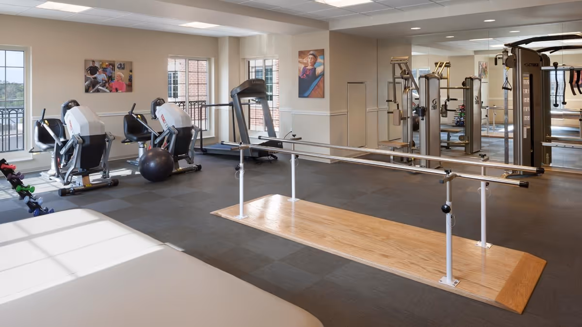 A well-lit senior living facility exercise room with various fitness equipment including stationary bikes, a treadmill, weight machines, a set of parallel bars for walking support, and dumbbells. The room has large windows, mirrors on one wall, and artwork hanging on the walls.