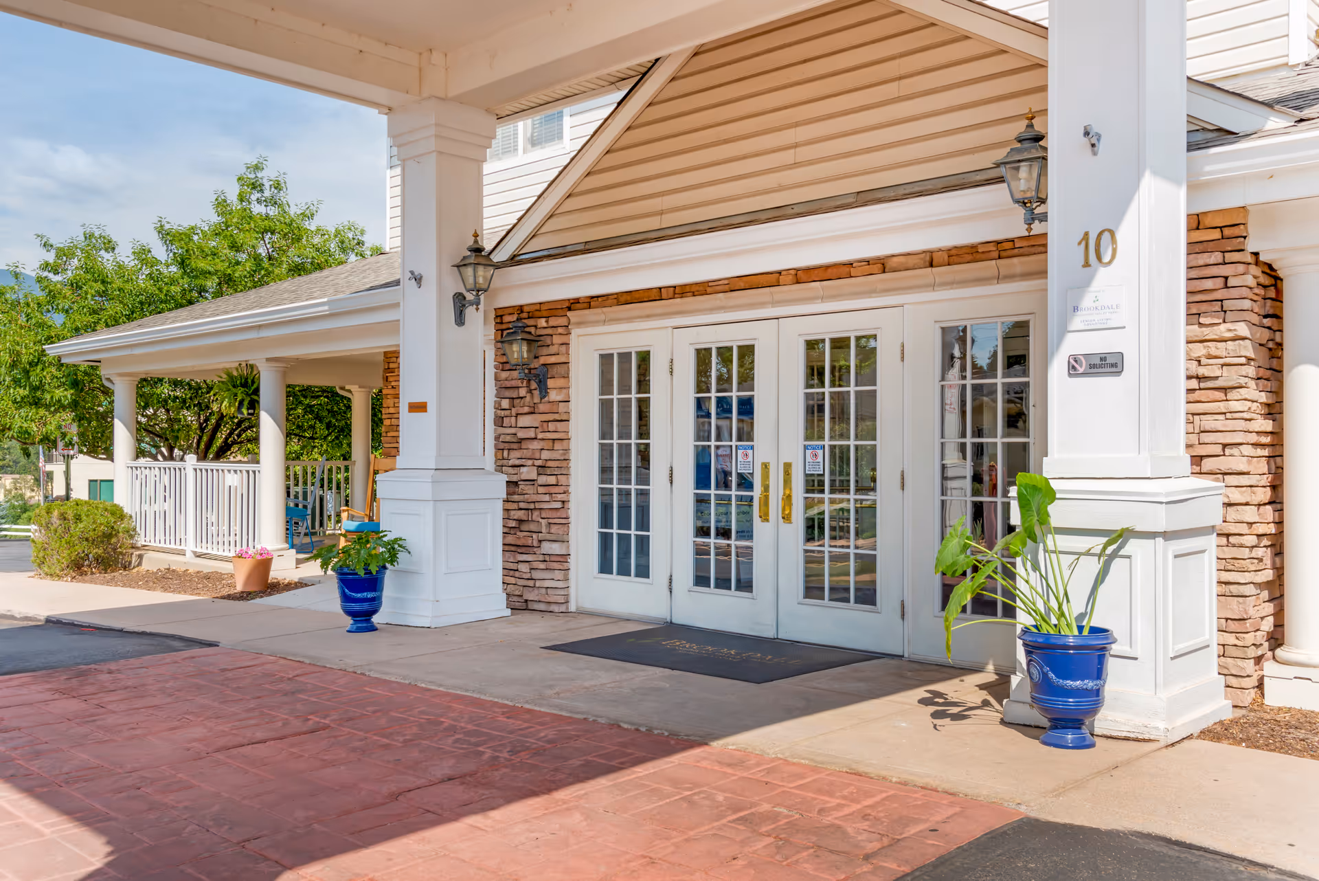 Entrance of Brookdale Monument Valley Park facility showing double glass doors with white frames, stone and siding exterior walls, two blue plant pots with green plants, white columns, and a covered driveway area.