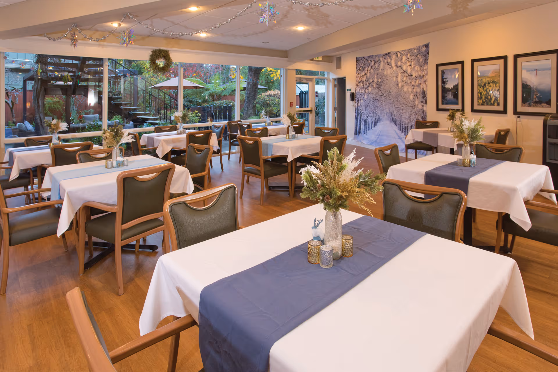 A dining room with multiple tables covered in white tablecloths and blue runners, each decorated with a vase of flowers and small candle holders. The room has wooden floors, green cushioned chairs, large windows showing an outdoor garden area, and framed pictures on the walls.