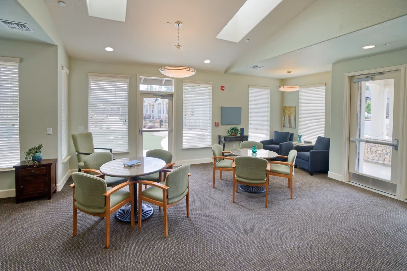 Bright communal lounge with round tables, green upholstered chairs, armchairs and large windows.