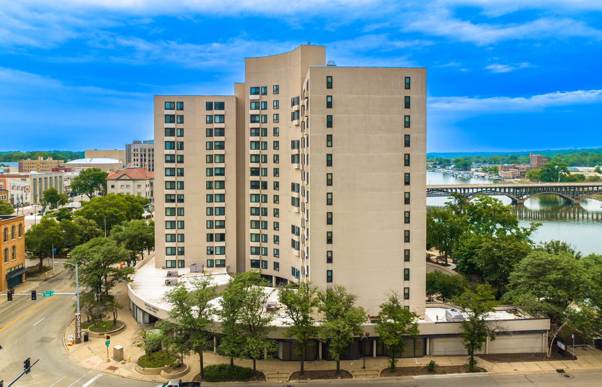 Exterior view of the Luther Center, a tall beige residential building beside a river and bridge in a downtown area.