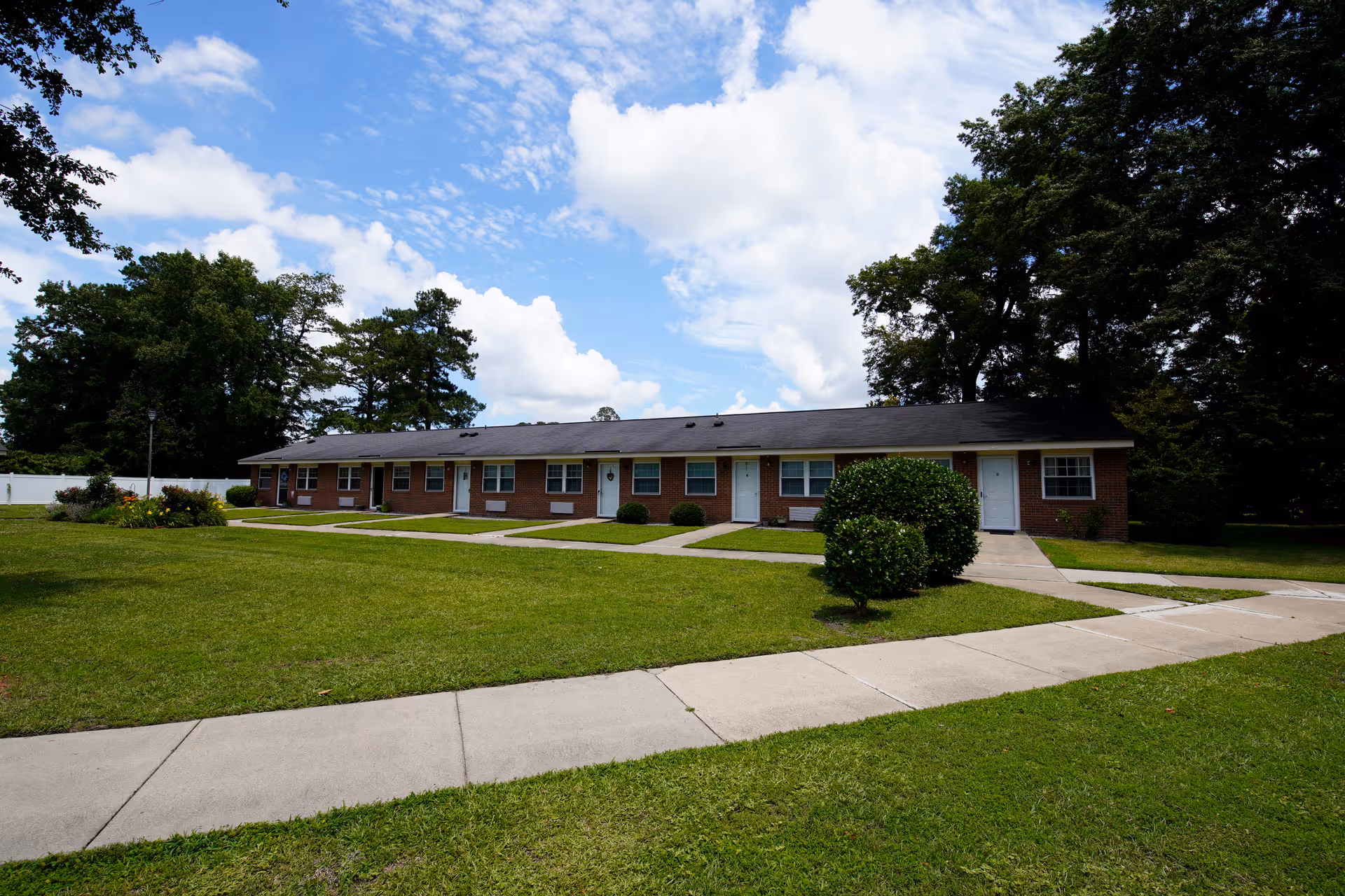 Single-story brick apartment building with a large grassy lawn, sidewalks, and trees beneath a partly cloudy sky.